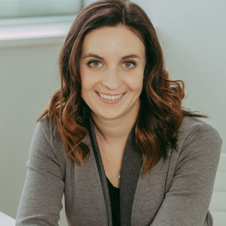 A woman with wavy brown hair, wearing a gray jacket and a black top, sits indoors, smiling at the camera.